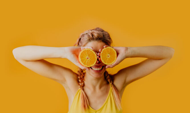 Photo of a playful young woman holding orange halves in front of her eyes; studio shot, isolated on an orange background